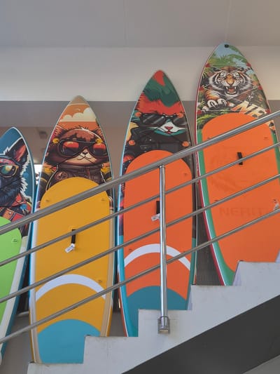 The image shows a display of stand-up paddleboards in a shop. The paddleboards are upright and leaning against a wall, with a metal railing in front of them. Each paddleboard features a different graphic design, including cartoon characters like a cat and a dog, and a tiger. The designs are colorful and elaborate. The shop appears to be indoors, possibly a retail store specializing in sporting goods. The lighting is bright, suggesting artificial illumination. The background is a plain, light-colored ceiling. There are no people visible in the image. The overall impression is a retail display of vibrant and artistically decorated paddleboards.