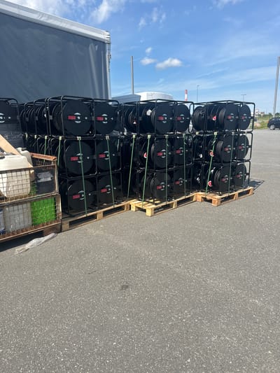 In Loo, Estonia, a stack of black cable reels is neatly arranged on wooden pallets in an outdoor setting. The reels are housed in black metal frames and secured with green strapping. To the left of the reels, a metal cage contains white and green containers. In the background, a large grey trailer is parked, and beyond that, industrial structures and a car are visible under a clear blue sky with scattered clouds. The ground is paved asphalt.