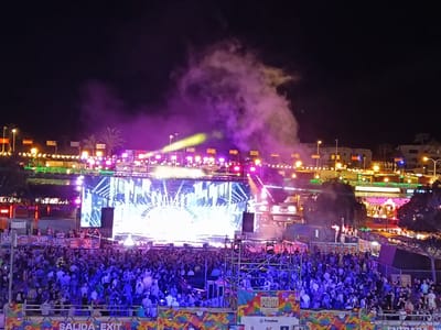 At Playa Del Inglés, Spain, a nighttime outdoor concert is in progress. A large crowd faces a brightly lit stage, with stage lights and smoke effects illuminating the performers. Buildings and flags line the background behind the stage. Fencing and signage separate the crowd from the stage area.