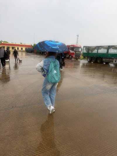 The image captures an outdoor scene on a rainy, overcast day, possibly in China. The primary subject is a person viewed from behind, walking away from the camera. This individual wears a light blue denim jacket over a grey hoodie, blue jeans, and white sneakers, and carries a light teal backpack with a white logo, possibly Adidas. They hold a distinctive blue umbrella patterned with a galaxy or stars and the word "PLANET" visible on its edge.

The ground is wet asphalt or concrete, showing numerous reflections from the rain, including those of the subjects and vehicles. In the mid-ground, a red semi-truck with "MANDAN" visible on its grille is parked. Behind it, a slender communication tower rises into the hazy, grey sky. To the right, part of a long, green semi-trailer is visible. Further in the background, towards the left, are low-rise buildings with yellow roofs and light-colored walls.

Other figures are present on the wet ground; one person in dark clothing pulls a small black rolling suitcase, and another dark-clad person stands further to the left, their reflections clearly visible in the wet surface. The overall atmosphere is dim and subdued due to the heavy overcast conditions, indicating inclement weather.