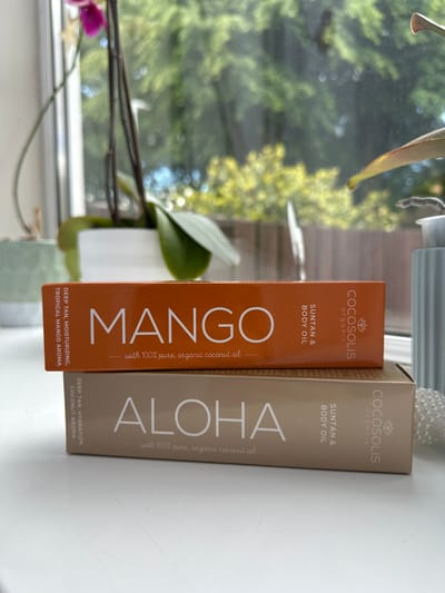 Two boxes of COCOSOLIS Organic Suntan & Body Oil are stacked on a white surface in front of a window. The top box is orange and features the word "MANGO" in large white letters. Text on the left side of the orange box reads "DEEP TAN, MOISTURIZING, TROPICAL MANGO AROMA". The bottom box is a light brown color and features the word "ALOHA" in large white letters. Text on the left side of the brown box reads "DEEP TAN, HYDRATION, COCONUT AROMA". Behind the boxes, an orchid plant is visible in a white pot, and beyond that, blurred green foliage suggests an outdoor environment. The scene is indoors, near a window, with natural light illuminating the objects. There are no people or animals visible in the image.