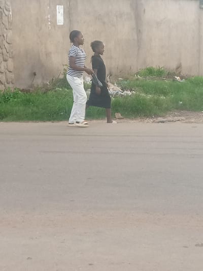 Two young females are walking on a paved surface adjacent to a grassy area with scattered debris. The female on the left is wearing a horizontally striped t-shirt and white trousers, with open-toed sandals. The female on the right is wearing a dark dress. Both are facing to the right. A wall made of stone or concrete is visible behind them, with a lighter colored, textured wall further back. The environment appears to be an outdoor urban or semi-urban setting. The location is Barikin Jirga Summa, Nigeria.