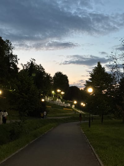The image depicts an outdoor scene at dusk in a park, likely in Vladikavkaz, Russia. A paved pathway curves through the park, bordered by grassy areas and trees. Several lampposts with bright, glowing lights illuminate the path and the surrounding foliage. In the distance, rows of white chairs are arranged, suggesting a gathering or event. The sky is a mixture of twilight hues, with dark clouds interspersed with lighter, warmer tones near the horizon. The overall atmosphere is serene and peaceful, with a hint of anticipation for the evening. There is no visible text in the image.