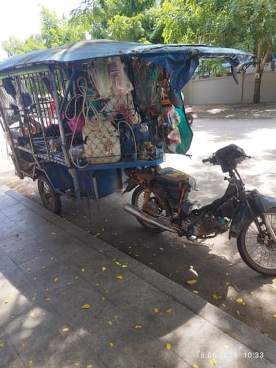 This media file depicts a modified motorcycle with a trailer attached, loaded with various items. The trailer is a blue, metal-framed cage covered with a tattered blue tarp, overflowing with what appear to be household goods, bags, and possibly recyclables. The motorcycle itself is dark-colored and appears well-used, with some items secured to the back.

The scene is set on a paved street next to a sidewalk covered in fallen yellow leaves. Sunlight filters through the trees overhead, casting dappled shadows on the ground. The overall impression is one of a mobile, utilitarian vehicle carrying a substantial amount of stored or collected belongings.

In the background, a gray concrete wall and lush green trees suggest an outdoor, possibly urban or semi-urban environment. A portion of another motorcycle or scooter is visible further down the street. The lighting suggests it is daytime, likely late morning given the shadow angles and timestamp.

The timestamp "18.06.2026 10:33" is visible in the bottom right corner, indicating the date and time the photo was taken. The location context is identified as Kampot, Cambodia. No people are visible in the image, and the scene conveys a sense of daily life and resourcefulness rather than any specific event. The condition of the vehicle and its cargo hints at it being used for transportation of goods or for a living.