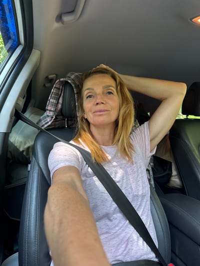 The image shows a woman sitting in the driver's seat of a car. She has blonde hair and is wearing a light pink t-shirt. Her right arm is raised, with her hand resting on the back of her head. A seatbelt is fastened across her body. The interior of the car appears to be dark leather, with a plaid shirt visible on the headrest of the passenger seat. The lighting suggests it is daytime, and the reflection on the window indicates an outdoor environment, possibly with trees. The image is a selfie, taken from the driver's perspective. There is no visible indication of the car being in Portovoe, Russia.