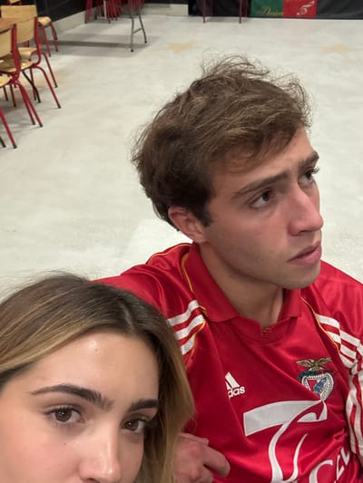 This indoor image captures two individuals in what appears to be a communal space within Décines-Charpieu, France, likely a community hall or cultural association.
In the foreground, partially visible on the lower left, is the upper portion of a female's face, featuring brown eyes, prominent eyebrows, and light brown hair with a central part. Her gaze is directed towards the viewer, suggesting she is holding the camera or is closest to it.
Behind her and to the right is a young adult male with messy light brown hair, his head turned upwards and to the right of the frame, not looking directly at the camera. He is wearing a red short-sleeved polo shirt with white stripes on the shoulders, a white Adidas logo on the right chest, and the S.L. Benfica crest on the left chest, with a white number '7' visible below it. His right hand rests near the bottom center of the frame, adjacent to the female's position.
The background features a light-colored, possibly concrete or tiled, floor. In the upper left quadrant, several chairs with red metal frames and light-colored seats are visible, some partially stacked. In the upper right, a red banner or decoration with white and possibly green details, including what appears to be a white musical note symbol and partial text, is displayed against a dark background.