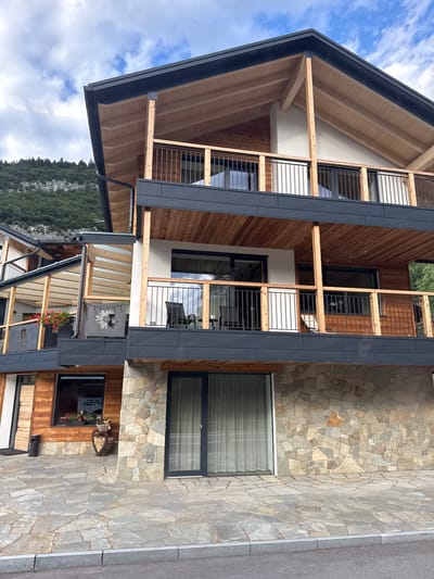 The image depicts the exterior of a multi-story building with a modern architectural style, incorporating natural materials like wood and stone. The building features several balconies with dark railings and wooden ceilings. The lower section of the building is clad in rough-hewn stone, contrasting with the polished wooden accents on the upper floors. Large glass doors and windows are visible. The surrounding environment includes a paved patio area with irregular stone tiles, a paved walkway with a curb, and a backdrop of a forested mountain under a cloudy sky. A decorative wooden heart-shaped sculpture filled with firewood is placed near the entrance. The building appears to be situated in a scenic, possibly mountainous, region. The sign visible in the window reads "12A".