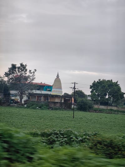 The image captures a view of a temple in Wadhona, India, on an overcast day. In the foreground, a vast green field of crops, possibly sunflowers or a similar low-lying plant, stretches across the landscape. The field is densely planted and appears to be slightly blurred, suggesting movement. To the right, a row of lush green bushes with some yellow flowers runs along the edge of the field.
In the midground, the temple building is visible. It features a prominent yellow and white spire, characteristic of Indian temple architecture. The temple is attached to a building with a red roof and blue-tinted windows, suggesting it might be part of a larger complex or a recently constructed addition. A large tree with spreading branches stands to the left of the temple, partially obscuring the building. Several other smaller structures and some greenery are also present in this area. A utility pole with electrical wires runs vertically through the midground, crossing the scene from left to right. Various agricultural equipment, including a tractor, are partially visible near the structures.
The background is dominated by a dull, cloudy sky, with some faint patches of lighter cloud suggesting the diffused light. The overall impression is of a rural or semi-rural setting in Wadhona, India, focused on the religious structure and the surrounding agricultural land. There are no people visible in the frame.