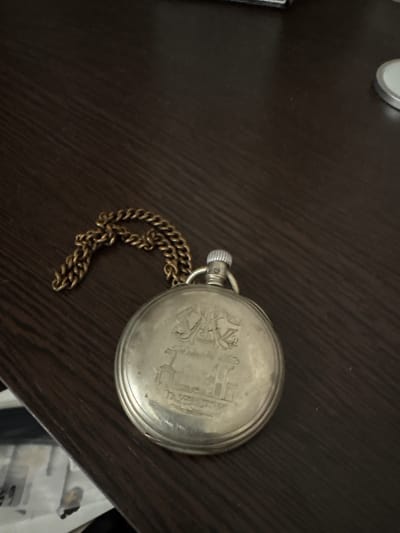 A silver-toned pocket watch with a gold-colored chain is resting on a dark brown wooden surface. The pocket watch is circular and appears to have an engraved design on its cover, which includes what looks like a ship and some lettering that is difficult to decipher. The chain is looped around the top of the watch. The lighting in the image is dim, casting soft shadows. The background is out of focus, but it appears to be a flat surface, possibly a table or desk. There are no people or animals visible in the image. The scene is static and appears to be an indoor setting. There is no identifiable information within the image that would allow for its location to be specified in Kinel'-Cherkasy, Russia.