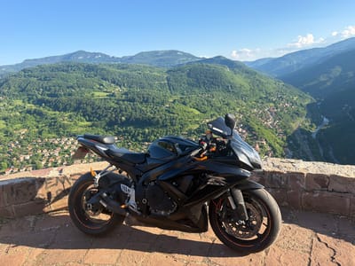 A black Suzuki GSX-R motorcycle is parked on a stone wall overlooking a valley. The motorcycle is in the foreground, angled slightly towards the viewer. The background shows a lush green valley with a small town nestled within it. Mountains rise in the distance under a clear blue sky. The scene is captured in Milanovo, Bulgaria, a location known for its scenic beauty and mountainous terrain.
