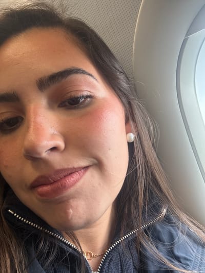 A close-up, tilted shot shows a young woman's face. She has brown hair, dark eyebrows, and fair skin. She is wearing a pearl earring. Her lips are a rosy color. A scar is visible on her chin. She's wearing a black jacket with a silver zipper and a gold necklace with a pendant shaped like a capital "D". Part of an airplane window is visible in the upper left corner of the frame. It is impossible to determine the specific location of the image in San Antonio based on the given information.