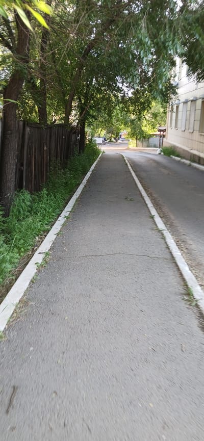 A paved walkway runs along a street in Chita, Russia. The walkway is bordered by a wooden fence on the left and a road on the right. Trees with green leaves overhang the walkway, casting shadows. A building with windows is visible on the right side of the road. The scene appears to be a residential or commercial area.