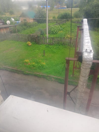 The image shows an outdoor scene in Asino, Russia, likely a residential area. In the foreground, a white surface, possibly a windowsill or railing, partially obscures the view. A large, insulated pipe, wrapped in reflective foil, is prominently featured on the right side of the image, supported by a red metal structure. Behind the pipe, a wooden fence encloses a grassy yard with some potted flowers. Beyond the fence, a small wooden house with a green roof and a satellite dish is visible, surrounded by vegetation. Further in the background, trees and distant buildings suggest a rural or semi-urban environment. The overall impression is one of a slightly overcast day, with evidence of rain or mist contributing to a hazy atmosphere. There are no people visible in the image.