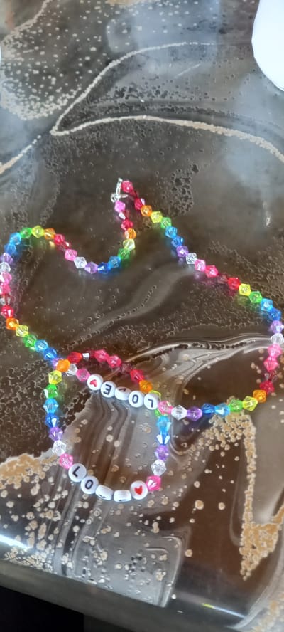 A close-up, overhead shot shows two beaded necklaces on a dark, textured surface. The necklaces are made of small, multi-colored faceted beads, predominantly in rainbow hues. One necklace is a single strand, while the other appears to be two strands joined together by a silver clasp. The larger necklace features white letter beads spelling out "LOVE," with a red heart bead also present. The smaller necklace consists solely of the colored beads. The background surface has a marbled or speckled pattern, with lighter colored specks and irregular darker lines. The lighting is somewhat dim, creating subtle shadows and reflections on the surface. The image is not geographically specific to Iskra, Bulgaria or any other city.