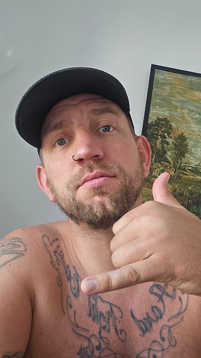 A man with a beard and a black cap is shown from a low angle, partially shirtless, with visible tattoos on his chest and shoulder. He is pointing down towards his chest with his right hand. He has blue eyes and a ring in his nose. A framed painting hangs on the wall behind him, which is otherwise plain white. There is no evidence to indicate the location is Los Alcázares, Spain.