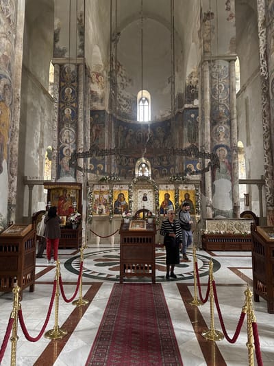 Here is a detailed description of the image:
The photograph captures the interior of Manasija Monastery, located in Despotovac Municipality, Serbia. The building has weathered and richly decorated walls covered with religious frescoes and artwork, which appear to have suffered from the ravages of time.
The floor is made of white marble with a geometric mosaic pattern. A long red runner with an ornate design lies on the floor, leading up to the altar area. Decorative brass stanchions and red velvet ropes create a pathway for visitors.
Several people are inside the monastery. One woman, wearing a striped top, is kneeling, and another individual is standing. On the other side, someone with red pants is kneeling near an icon. The subjects appear to be engaged in prayer or observation of the religious art.
The interior features an altar adorned with religious icons and flowers. The light in the room filters through the windows, creating a contrast between the illuminated and shadowed areas.
The composition of the image is vertical, providing a broad view of the monastery's interior.