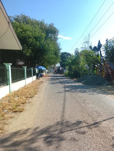 The image depicts a sunlit, paved road in Manyang Lancok, Indonesia. On the left side of the road, a black metal fence with light green posts borders a property with dry grass and some scattered litter in the foreground. A white-roofed building partially juts into the frame from the top left. Beyond the fence, lush green trees create a dense canopy. Further down the road on the left, a blue umbrella shades a small stall, with a person visible next to it.

The road itself is a granular asphalt surface, with tire tracks and shadows of overhead elements cast across it. On the right side of the road, a large pile of grey gravel sits beside a banana tree and other foliage. Further down the road, a tractor is parked. In the distance, there are more trees, houses, and power lines stretching across the bright blue sky, with a single white cloud visible. Two motorcycles are seen on the road in the distance, one approaching and one moving away. The overall scene suggests a typical village or suburban environment in a tropical climate.