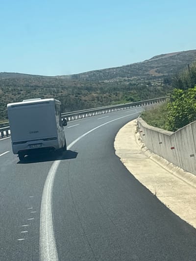 An aerial shot shows a white camper van driving on a highway that curves to the right. The highway is paved with asphalt and has a white lane marker. The camper van is positioned in the left lane, with its rear facing the camera. The background consists of rolling, arid hills covered in sparse vegetation, under a clear blue sky. A concrete barrier with red reflectors lines the right side of the highway. The scene is set in Avra, Greece.