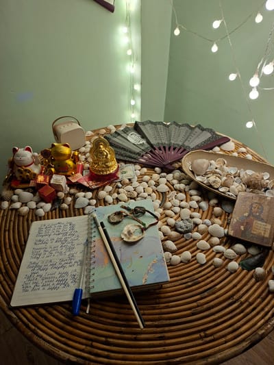 The image shows a close-up view of a round, woven table with various objects placed on it, set against a pale green wall decorated with string lights. The table's surface is covered with numerous seashells.
Prominently displayed on the table are several decorative items: a white and red Maneki-neko (beckoning cat) figurine, a golden cat figurine, a golden Buddha statue, and a folded fan with East Asian script. Among these are several US dollar bills and small, red and gold boxes. A small, light-colored portable speaker is also visible.
In the foreground, a spiral-bound notebook with a blue and green cover is open. The notebook contains handwritten text in English, a pencil, a pen, and some small, circular objects that appear to be shells or lids. Also on the table are a small, square wooden icon of a religious figure, and a shallow oval dish filled with more seashells.
The overall impression is one of a curated collection of decorative and symbolic items, possibly for personal reflection or display. There is no indication of the specific location within Sihanoukville, Cambodia, beyond what can be inferred from the general decor. The scene appears to be indoors, with artificial lighting from the string lights. No people are visible.
