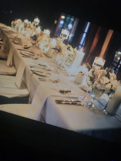 A long table arrangement features candles and flowers in glass containers. The table is covered with a white tablecloth. Silverware and small sprigs of greenery are placed beside each setting. The background shows hints of an indoor setting with beams or windows, suggesting a wedding reception or formal event. The image is captured at an angle, with some parts out of focus, creating a soft, romantic ambiance.