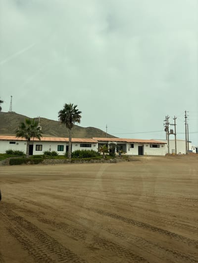 The image depicts a low-rise building with a terracotta tiled roof, painted white, situated in a dusty, arid environment. Two palm trees are prominently featured in front of the building. In the background, a hilly, barren landscape is visible, topped with a tall communications tower on the left side. On the right side of the image, electrical poles and a large white cylindrical tank suggest industrial or utility infrastructure. The foreground consists of a dirt or sand area with visible tire tracks, indicating vehicular activity. The sky is overcast. The scene is located in the city of San Antonio, Peru.