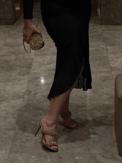 A person's legs and hands are visible in this image, which appears to be taken indoors on a marbled floor. The person is wearing a black dress, gold sparkling heels, and holding a matching gold sparkling clutch. The lighting is dim, creating shadows on the floor. The location is not discernible from the image itself, and therefore, cannot be confirmed as Cebu City, Philippines.
