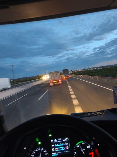The media file depicts a view from inside a truck cabin, looking out at a highway at dusk. The subjects include several trucks on the road, with one in the foreground displaying its rear lights. The scene is set on a highway with multiple lanes, separated by a concrete barrier and illuminated by streetlights and the headlights of vehicles.

The time of day is clearly dusk, indicated by the twilight sky with a mix of blue and gray clouds, and the artificial lighting on the road and in the distance. The weather appears to be clear, with no visible precipitation. The location is identified as Picassent, Spain, which is suggested by the presence of a highway and the surrounding landscape with some vegetation and buildings visible in the distance.

Notable details include the dashboard of the truck, which shows various readings, including temperature (21°C), a digital display with numbers like "11" and "34," and a speedometer with a needle pointing towards the lower end. The steering wheel is also visible, framing the view. The overall mood is that of a journey in progress, with the approaching darkness and the illuminated highway creating a sense of movement and transition. The activity is simply driving on the highway.

The visible text from the OCR includes "OS," "60," "90," "3.12," "Q," "34," "11," "64," "15," "10," "20," "100," "5," "255," "L," "1-," and "2," likely representing units and readings from the truck's dashboard.