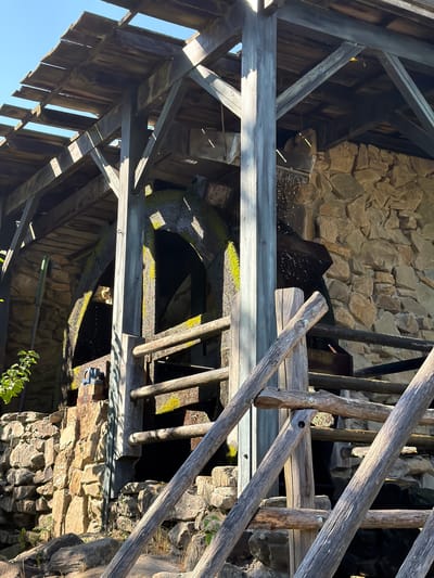 This is a photograph of an old watermill in Heijen, Netherlands, under a clear blue sky. The mill is constructed of wood and stone, with a large water wheel partially visible. The wooden structure is weathered and shows signs of age, with planks of the roof missing in places, allowing sunlight to filter through. The water wheel itself is also aged, with moss growing on its surface. Water is seen cascading from the wheel and splashing onto the stones below. A rough-hewn wooden staircase or walkway leads up to the mill. The background is a stone wall, and the surrounding area appears to be rustic and natural, with some greenery visible.
