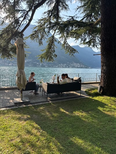This image shows a scenic outdoor seating area at a lakeside establishment in Pognana Lario, Italy. The scene is set under the dappled shade of large evergreen trees, with a panoramic view of a lake and distant mountains dotted with houses. In the foreground, a lush green lawn stretches out, catching the warm light of the sun.
The seating area features two dark-colored modern couches arranged around a small table. On the couches, three individuals are visible. One person, on the left couch, faces away from the camera and appears to be looking out at the lake. Two other individuals are seated on the right couch, facing each other and engaging in conversation or enjoying drinks. Their interaction suggests a relaxed social gathering.
On the table, several items are present, including glasses, bottles, and what appear to be drinks. An umbrella, in a neutral beige color, is positioned to the left of the couches, providing shade. The ground beneath the seating area is paved with cobblestones, and a metal railing separates it from the lakefront. The lake itself is a vibrant blue, with gentle ripples on its surface. The background showcases the mountainous landscape of Pognana Lario, with buildings clustered along the slopes, indicating a populated area. The overall atmosphere is peaceful and inviting, highlighting a typical summer day in this Italian lakeside town.