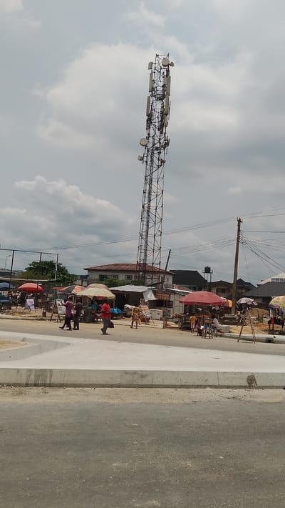 The image depicts a scene in Effurun, Nigeria. A tall, gray telecommunications tower dominates the view, rising high above the low-rise buildings and market stalls that line the street. Several people are walking across what appears to be a newly constructed or renovated section of pavement. Market stalls with colorful umbrellas and goods are visible in the background, suggesting a commercial or trading area. The sky is overcast with gray clouds. Overhead power lines run parallel to the street.