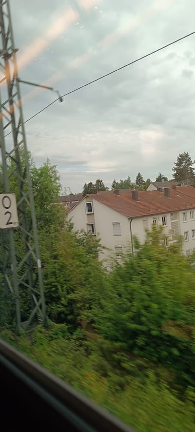 An exterior, medium shot captured from a moving vehicle shows a residential area in Waiblingen, Germany. In the foreground, a metal pylon with the number "02" is visible on the left. A overhead power line runs diagonally across the frame. Behind the pylon, lush green trees obscure much of the view. In the midground, a white multi-story building with red-tiled roofs and numerous windows stands prominently. The sky is overcast with light clouds. The motion blur indicates the photograph was taken from a moving train or car.
