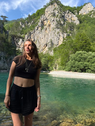 A young woman stands in the shallow, clear, turquoise water of a river. She is wearing a black crop top and black shorts. Her long, brown hair is down, and she is looking up and to her left. The riverbank behind her is covered in white pebbles. In the background, steep, rocky mountains covered in green trees rise towards a blue sky with scattered white clouds. The scene is set in the city of Bistrica, Montenegro, a natural environment characterized by its pristine river and dramatic mountainous terrain.