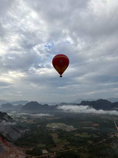 A red and yellow hot air balloon is visible in the sky over a mountainous, rural landscape. The balloon is positioned in the upper center of the image. Below the balloon, a valley is visible with lush green trees, agricultural fields, and a body of water. In the foreground to the left, a rocky incline with a quarry is present. The sky is filled with dramatic, cloudy formations. The location is identified as Hong Ngern Village, Laos.