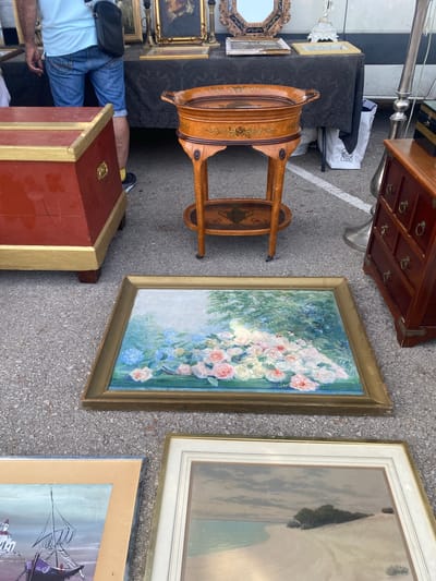 The image depicts an outdoor market stall, likely in San Luis De Sabinillas, Spain, given the context provided. In the foreground, laid flat on the asphalt, are two framed paintings. The upper painting features a floral arrangement with pink and white roses amidst blue hydrangeas and green foliage, framed in gold. Below it, another framed painting shows a boat with purple sails docked near white buildings by the water.

Behind the paintings, a red wooden chest with gold trim is visible on the left. To the right of the chest, a person wearing blue jeans and a light blue t-shirt stands with a black bag slung over their shoulder. Further to the right is a wooden stand with two tiers, both decorated with an inlaid design of birds and floral patterns; the upper tier holds a circular wooden tray. Beyond this, a dark brown wooden cabinet with multiple drawers and brass hardware is positioned. In the background, a dark tablecloth covers a table displaying various framed objects and decorative items, including a portrait and a mirror. A white shopping bag with a logo is tucked beneath the table. The overall scene suggests a vendor selling antiques or artwork at an outdoor market.