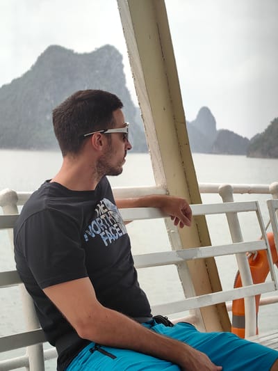 A man sits on the railing of a boat, looking out at the water and rock formations. He is wearing a black t-shirt, blue shorts, and sunglasses. The background shows a misty sky, the sea, and several rocky islands. The boat's railing is white, and an orange life preserver is visible on the side of the boat. The location is in the city of Hải Phòng, Vietnam.