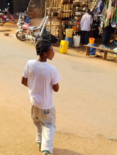 The image depicts a daylight street scene in Enugwu-Ukwu, Nigeria. In the foreground, a young male with dark skin and braided hair wears a white short-sleeved t-shirt, light-colored loose-fitting pants, and sandals with green and white striped socks. He walks on a reddish-brown dirt shoulder, facing away from the camera but looking back over his right shoulder. His form casts a shadow to his left. The adjacent road is paved and light tan.
On the left side of the road, two motorcycles are parked on the dirt shoulder; one is a red motorcycle with silver accents and a clear windshield, positioned upright on a kickstand. A dark-colored, weathered building is visible in the far left background.
On the right side of the paved road, a market stall composed of wooden shelving displays various items, including hanging colorful plastic and metallic objects. A male individual, dressed in a light-colored long-sleeved shirt and dark trousers, stands with his back to the camera, positioned at this stall. Numerous plastic containers, specifically yellow jerry cans, blue cylindrical barrels, white buckets, and orange containers, are arranged on the ground and stacked near the stall. A light-brown wooden bench is situated horizontally in front of the stall. The scene is illuminated by natural light, producing distinct shadows.