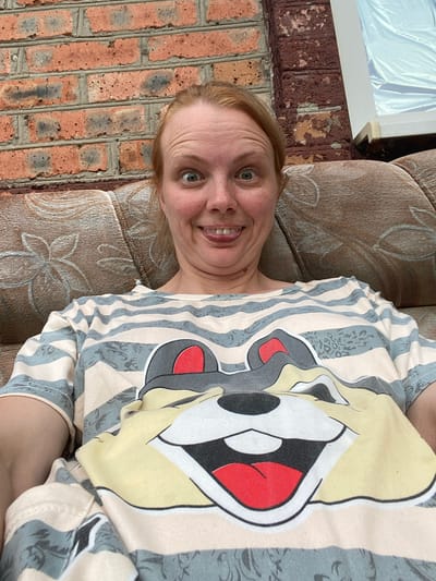 A woman with red hair and a striped t-shirt with a cartoon character print is sitting on a couch in front of a brick wall. She is sticking her tongue out and her eyes are wide open. The couch is brown with a floral pattern. The brick wall is made of orange bricks with some grey mortar. There is a window to the right of the image covered with a white sheet.