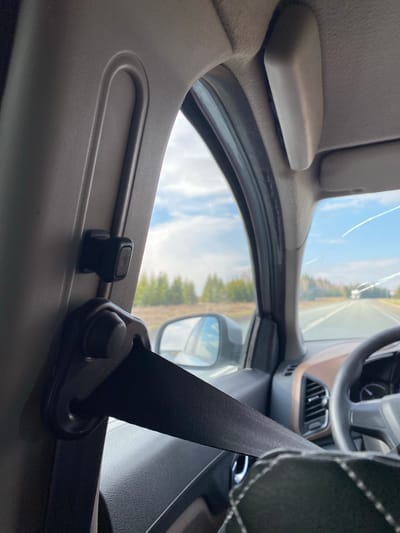 The image depicts the interior of a car, specifically the area around the driver's side B-pillar. A black seatbelt is prominently visible, with its height adjustment mechanism attached to the B-pillar. Outside the car window, a road and foliage are visible under a partly cloudy sky. The steering wheel and dashboard are partially visible, suggesting the photo was taken from the passenger seat or the back. The exact location in the city of Bal'zyashur, Russia cannot be determined from this image alone. There are no people visible in the image.