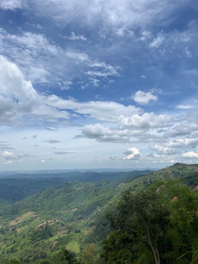 The image displays a panoramic view of a lush, mountainous landscape under a partly cloudy sky. The foreground features dense green foliage, including trees and vegetation, with a visible tree trunk and branches dominating the right side of the frame. The midground reveals rolling hills covered in a mosaic of green vegetation, with some areas showing signs of cultivation or sparse tree cover. The background consists of multiple layers of receding mountains, fading into a hazy horizon. The sky is a vibrant blue, punctuated by scattered white and grey clouds, some of which appear wispy and others more substantial. The overall impression is one of natural beauty and expansive, undeveloped terrain. The location is identified as Son Dien city, Vietnam.