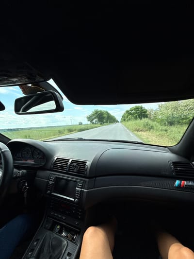 The view from inside a car shows a road stretching ahead with trees lining either side. The car's dashboard, vents, rearview mirror, and steering wheel are visible. A person's leg is visible on the right side of the frame. The location, Karavelovo, Bulgaria, cannot be confirmed based on the image alone.
