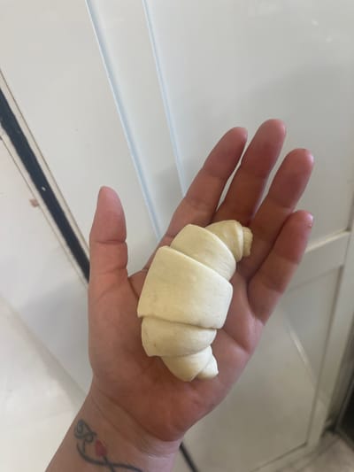 A human hand is holding a raw croissant. The hand is open, palm up, and the croissant is resting on it. The croissant is light yellow and appears to be made of dough. The hand has a tattoo on the wrist. The background is a white, tiled surface. The image is taken from a top-down perspective. There is no information about the location in Vallo della Lucania, Italy.