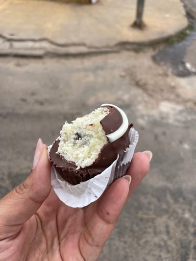 A close-up, eye-level photograph depicts a human hand holding a partially consumed, chocolate-covered confection in an outdoor, urban environment identified as Anápolis, Brazil. The confection, held in a white fluted paper cup, has a dark brown chocolate shell. A bite has been taken from its upper left portion, revealing a white, shredded coconut filling with small, dark inclusions. A white chocolate swirl or crescent decoration is visible on the top right of the confection. The hand is light brown in skin tone, with visible thumb, index, and middle fingers supporting the treat. The fingernails are short and unpolished. The background is out of focus, consisting of a textured, grey surface consistent with concrete or asphalt pavement, showing indistinct lines and variations in tone. A blurry, light-colored vertical pole or post is faintly visible in the upper right background. The lighting is natural daylight, indicating an outdoor setting.
