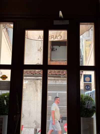 A man walks past a storefront in Sarajevo, Bosnia and Herzegovina. The storefront has a large wooden door with multiple glass panes. Through the glass, awnings, tiled roofs, and building facades are visible. The man is wearing a light-colored t-shirt and shorts, sunglasses, and is walking from left to right. He appears to be in his 40s or 50s. On the glass panes are various stickers and signs, including what appears to be a "Booking.com" rating and a warning sign with a skull and crossbones. The overall scene is bright and sunny, indicating it is daytime.