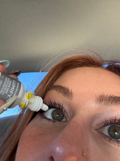 The image shows a person applying Lumify eye drops. The person is holding a bottle of Lumify in their hand, and the dropper is positioned near their left eye. The person has red hair and a nose piercing. The label on the bottle reads "BAUSCH+LOMB LUMIFY BRIMONIDINE TARTRATE OPHTHALMIC SOLUTION 0.025% REDNESS RELIEVER EYE DROPS Sterile 0.25 FL".

There is insufficient information to determine whether the image was taken in the city of Coachella, United States of America.