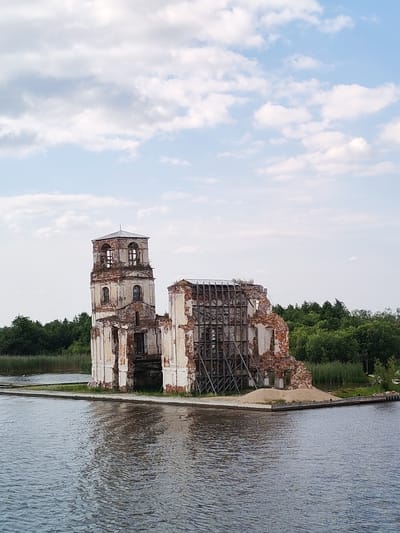 An image of the Church of the Nativity of the Theotokos in Krokhino, Russia, shows the partially ruined structure situated on a small island in a body of water. The church has a tall bell tower on the left, with a partially collapsed roof and exposed brickwork. The main body of the church to the right is heavily damaged, with large sections of walls missing, revealing internal scaffolding. The building appears to be made of brick and stone, with white plaster visible in some areas. A pile of sand is present on the ground next to the damaged section of the church. The church is surrounded by water, and a dense green treeline is visible in the background. The sky above is partly cloudy, with white and light blue clouds. The overall scene depicts a historical landmark in a state of disrepair, possibly undergoing restoration. There are no people or animals visible in the image.