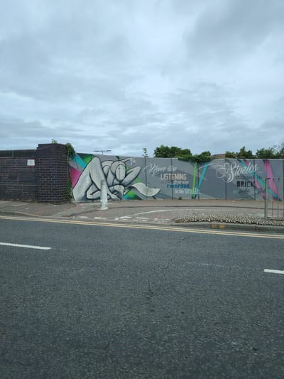 This is an outdoor, daytime photograph taken in Peterborough, United Kingdom, depicting a section of Gladstone Street. The scene features a long, low concrete wall adorned with a multi-part graffiti mural under an overcast sky. The mural includes a stylized grey figure in a reclining pose against a background of diagonal pink, green, and blue stripes. Adjacent to the figure, text in white and green reads: "Never stop LISTENING. The tales that are recorded in the Walls." Further to the right, another section of the mural displays ornate white text stating: "Stories trapped in BRICK that wait to be told." To the left of the mural, a dark red-brown brick wall forms a corner, bearing a small white rectangular sign and green ivy climbing its surface. In the foreground, an asphalt road with double solid yellow lines and a single solid white line runs horizontally. A concrete pavement borders the road, on which a white concrete bollard stands in front of the mural's figure, and a patch of small, light grey decorative stones is visible further along the pavement. The sky is uniformly grey and heavily overcast, with no direct sunlight. No people or vehicles are present in the scene.