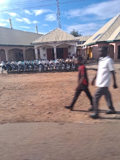 The image displays an urban street scene in Beli, Nigeria, under a blue sky with scattered white clouds. The foreground consists of a concrete strip transitioning into a wide expanse of reddish-brown dirt. Two individuals are captured in motion blur, walking from right to left across the dirt; the person on the left wears a dark top and dark pants, while the person on the right wears a white top and dark pants.
In the mid-ground, a line of approximately 20-30 motorcycles, mostly silver and white, are parked directly in front of a single-story building. This building features a light-colored pitched roof, arched entrances, and "Honda" logos affixed to its facade. To the left and slightly behind this structure is a larger building with a similar light-colored pitched roof and two minarets, suggestive of a mosque, displaying a circular design on its visible wall. To the right of the central building, another single-story building with a light-colored pitched roof and reddish-brown walls is visible, with a faint human figure in one of its doorways. A tall, lattice-style communication tower with multiple antennas stands prominently in the background, positioned to the right of a visible green tree. Overhead wires are visible across the top of the frame.
