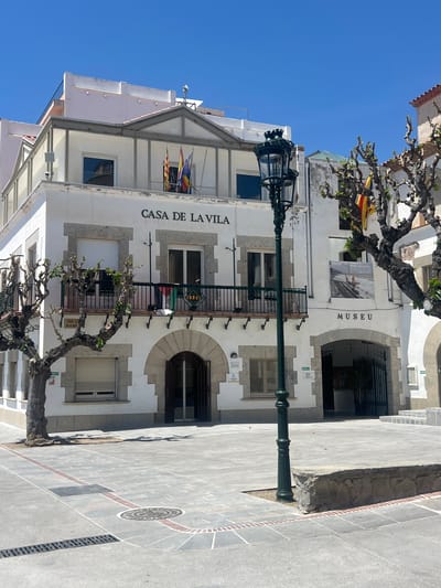 A daytime street scene in Sant Pol de Mar, Spain, is depicted under a clear blue sky. The image is oriented vertically (rotated 90 degrees clockwise). The main subjects are two white buildings with grey stone accents and wrought-iron balconies, facing a paved street.
One prominent building is identified by the text "CASA DE LA VILA" (House of the Village/Town Hall) above its ground-floor windows. Flags, including the Spanish and Catalan flags, are flying from its upper floor. On the ground level, a sign also reads "PLACA DE LA VILA" (Village Square).
Adjacent to or part of this structure is another section labeled "MUSEU" (Museum) with an arched entrance. A poster is displayed on the museum wall, featuring text that reads "PERE SAULEDA DONAR A VEURE SANT POL".
In the foreground and mid-ground, a tall, ornate dark green lamppost stands prominently. Several trees with intricate, mostly bare branches and some emerging green leaves are visible, framing parts of the buildings. The street below is paved with grey and reddish-brown tiles, featuring a circular manhole cover and a patterned design. No people or active events are visible, suggesting a quiet moment. The bright sunlight indicates it is daytime.