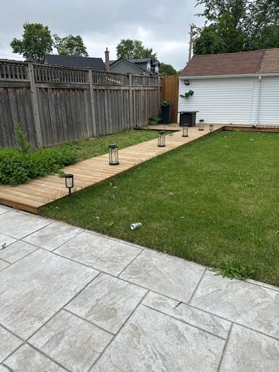 This image captures a backyard scene in Mississauga, Canada, under an overcast sky, suggesting it is likely daytime, possibly late afternoon or morning. The scene depicts a well-maintained backyard with distinct areas: a wooden deck, a grassy lawn, and a paved patio.
The wooden deck runs along the side of a white-sided house, featuring a dark-colored patio furniture set consisting of two chairs and a small table. Several modern, dark-colored outdoor lanterns are strategically placed along the edge of the deck, adding to the ambiance. Adjacent to the deck is a lush green lawn dotted with a few fallen leaves.
Further to the right, a paved patio area is visible, composed of large, light-colored square tiles with a stone-like texture. A discarded aluminum can rests on the grass near the edge of the patio.
The background is dominated by a tall, weathered wooden fence that separates this property from neighboring houses and greenery. Trees are visible, adding to the suburban feel of the location. The architecture of the nearby houses, with their sloped roofs and siding, are typical of residential areas in Canada. The overall impression is one of a functional and pleasant outdoor living space, likely for relaxation and entertaining. There is no visible text in the image.