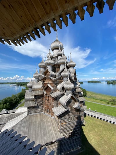 The image displays a detailed view of the Kizhi Pogost, a historical architectural ensemble located on Kizhi Island in Karelia, Russia, near the city of Boyarshchina. The primary subject is the Church of the Transfiguration, a magnificent wooden church characterized by its numerous domes and intricate wooden construction. The church is situated within a grassy, green landscape adjacent to a body of water, likely Lake Onega, with islands visible in the distance. The sky is a bright blue with scattered white clouds. In the foreground, there is a wooden structure, possibly a viewing platform or part of another building, with distinctive carved wooden elements. Two individuals are visible walking on a wooden pathway to the right of the church, suggesting the area is a tourist or historical site. The image is taken from an elevated perspective, looking down and across the church.